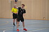 Håndballspiller i svart drakt kaster ballen i en innendørs hall med dommer bak.