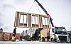 Kranbil løfter stort veggelement i tre mens flere arbeidere sikrer på byggeplassen.