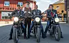 Tre motorsykler med førere står parkert foran et rødt trehus på en gate.