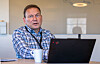 En mann sitter ved et skrivebord med laptop og kaffekopp i et kontor med glassvegg.