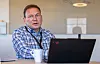 En mann sitter ved et skrivebord med laptop og kaffekopp i et kontor med glassvegg.
