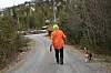 Person i oransje jakke går på grusvei med hund i skogkledd område.