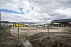 Anleggsområde bak metallgjerder med grus, maskiner og bygninger under en skyet himmel.
