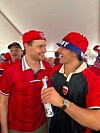 To menn i røde supporterklær og capser står tett sammen og smiler i et telt med flere mennesker i bakgrunnen.