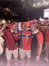 Tre personer i røde norske trøyer holder et norsk flagg i en innendørs arena med publikum og lys i bakgrunnen.