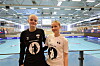 To ungdommer står smilende i en håndballhall foran en blå spilleflate og tribuner.