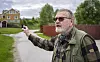 En eldre mann med solbriller peker mot et område ved en grusvei, med hus og gjerde i bakgrunnen.