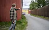En mann står ved en rød bygning og peker ned en smal vei med gjerder og fargerike hus i bakgrunnen.