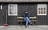 En mann sitter på en benk foran et svart trehus med to vinduer og en rød kost.