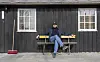 En mann sitter på en benk foran et svart trehus med to vinduer og en rød kost.