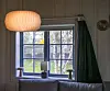 En liten stue med sofa, vindu, gardiner og hengende taklampe i et tømmerhus.