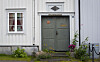 Inngangsparti i en lys trebygning med grå dobbeltdør, blomsterbed og skilt over døren.