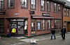 To personer går forbi en rød trebygning med butikk, skilt og utstilt jakke.