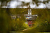 Hvit kirke og rødt trehus omgitt av høstskog, sett gjennom uskarpe greiner.