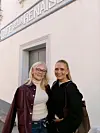 To kvinner poserer foran en kaféinngang med skilt på en lys fasade.