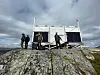 Personer ved et solcelleanlegg på en steinete fjellrygg under mørke skyer.