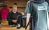 Håndverker arbeider ved en benk i et treinnredet verksted, delvis skjult av en person i grønn T-skjorte.