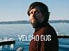 Skjeggete mann i mønstret genser ved vannet med fjell i bakgrunnen og teksten «VELCRO DOG».