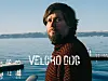 Skjeggete mann i mønstret genser ved vannet med fjell i bakgrunnen og teksten «VELCRO DOG».